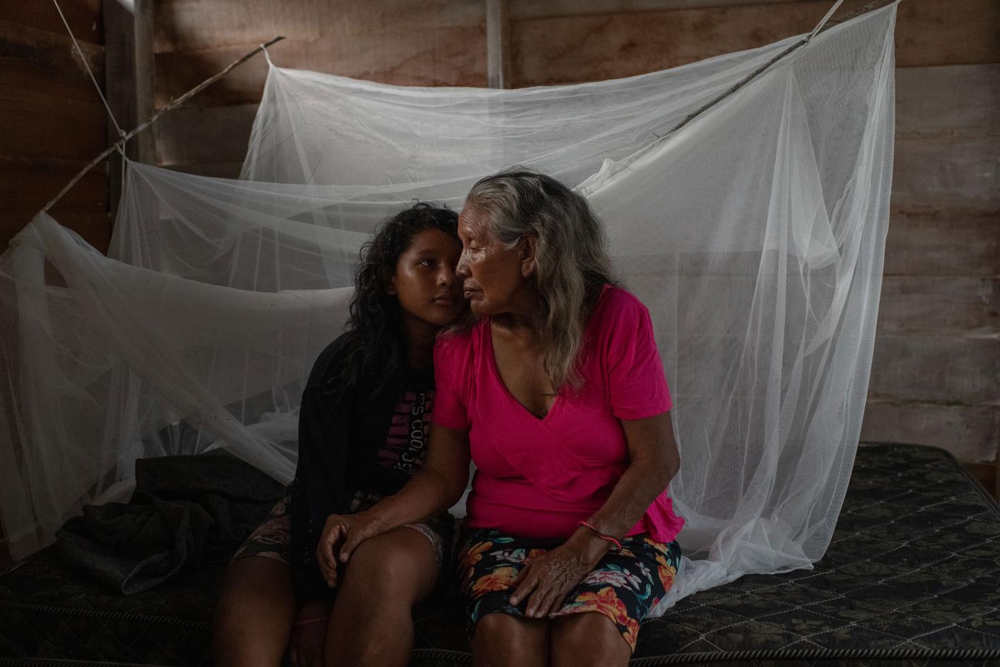 A granddaughter and grandmother sit close to one another in front of hanging mosquito nets