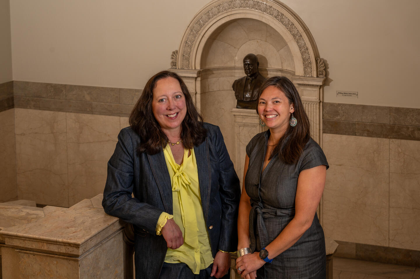 Two women, Associate Dean Anne Seymour and Associate Director of Informationist Services Beth Whipple, pose in the Welch Medical Library in front of a bronze bust