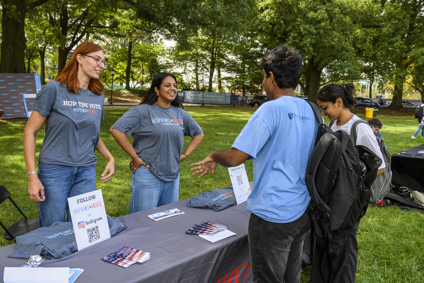 Two adults wearing Hopkins Votes shirt stand behind an outdoor voter registration booth. Two college students talk to them from across the booth.