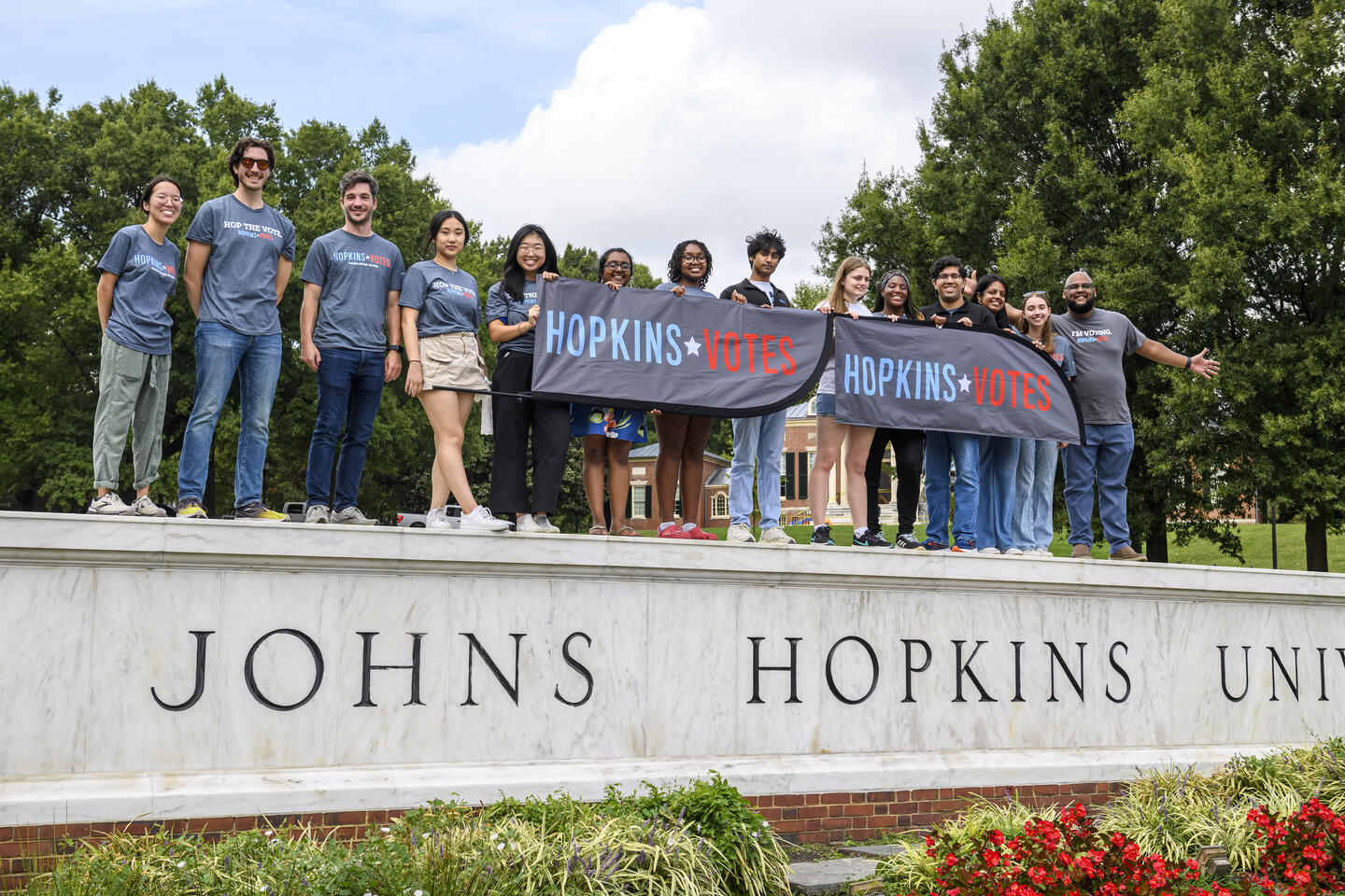 A group of Hopkins Votes volunteers wearing matching shirts and holding up Hopkins Votes banners stand atop the Johns Hopkins University sign.