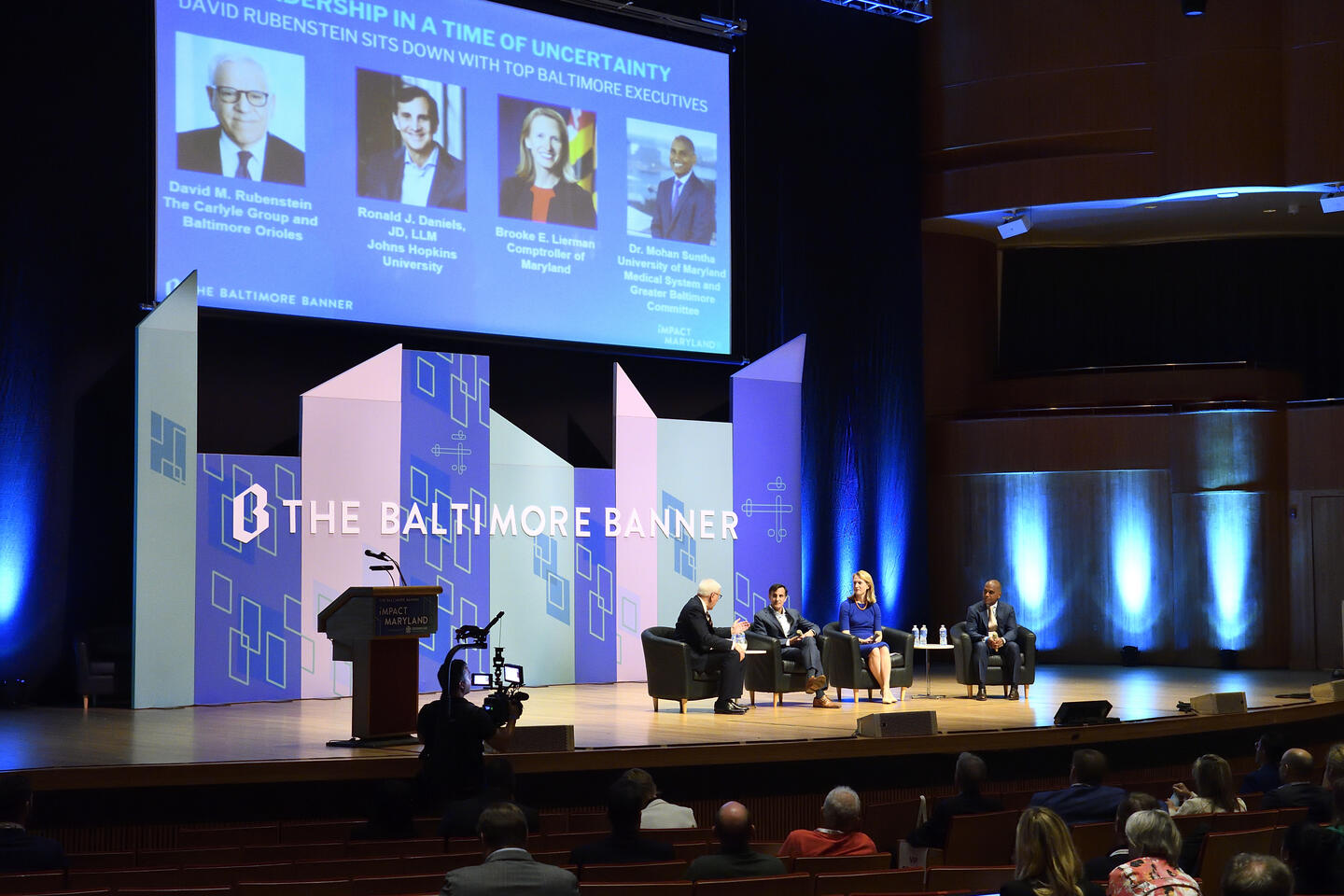 Four people sit on chairs onstage. Behind them is a sign that reads The Baltimore Banner, as well as a projector with their names and faces. The names on the projector read: David M. Rubenstein, Ronald J. Daniels, Brooke E. Lierman, and Mohan Suntha.