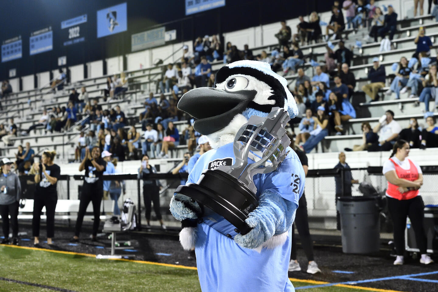 Jay the Blue Jay mascot carries a large silver trophy across Homewood Field. Behind them, people are sitting in the field's stands.