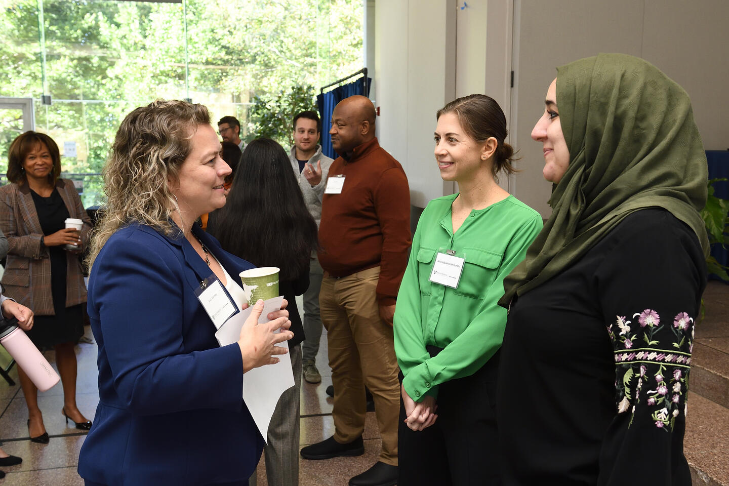 Three adults talk to each other while standing in a circle. Behind them, others do the same in their own groups.