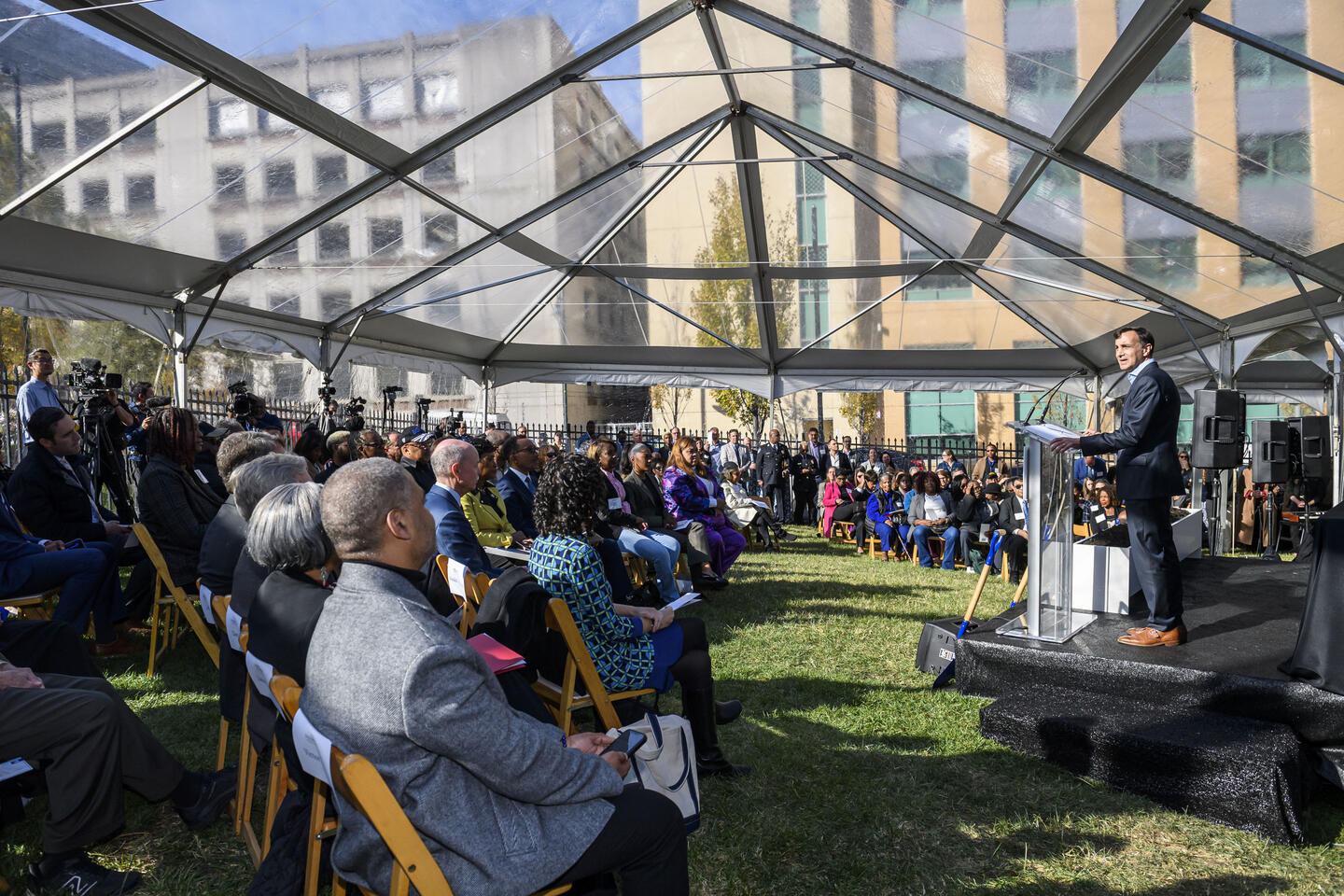 A man stands at a podium and addresses a large crowd beneath a clear tent