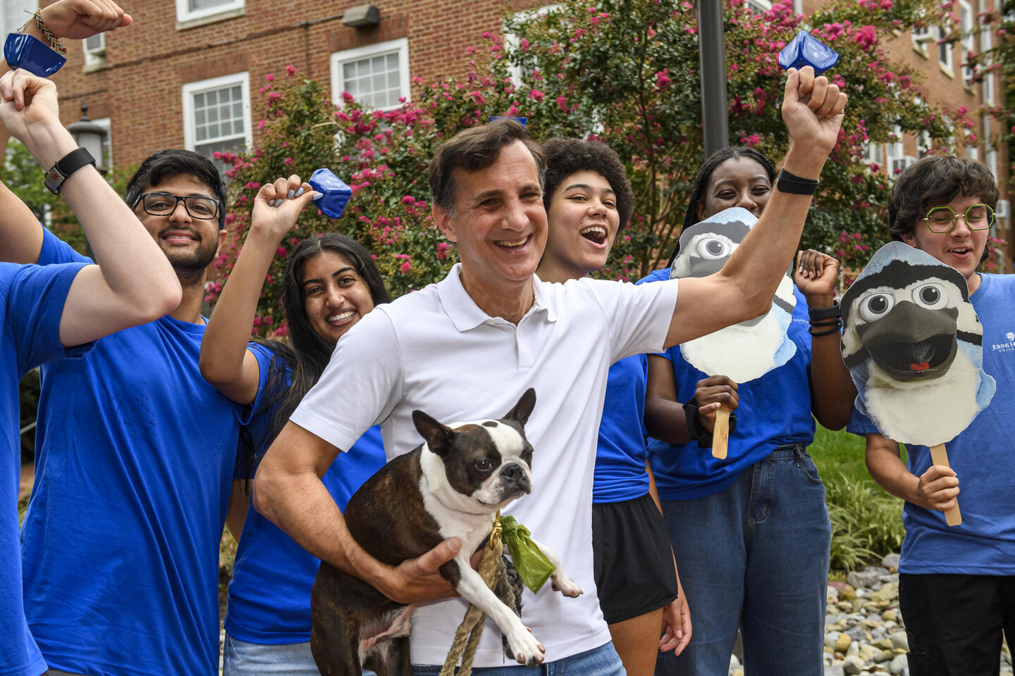 Ron Daniels waves a bell in the air while holding a small dog. Behind him, college students in blue shirts cheer.