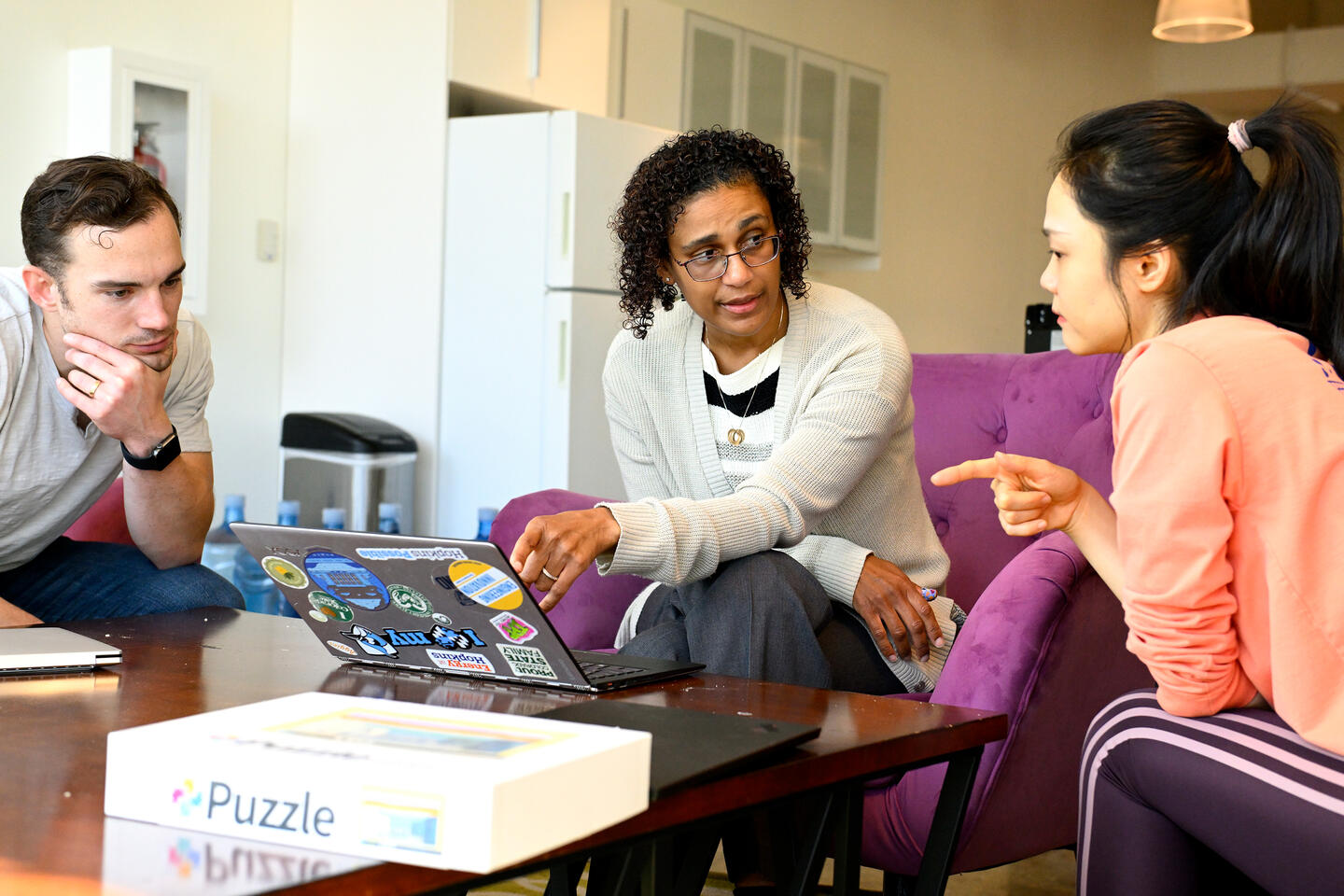 Three people sit and converse around a laptop, which is decorated with several colorful stickers. A box that reads 