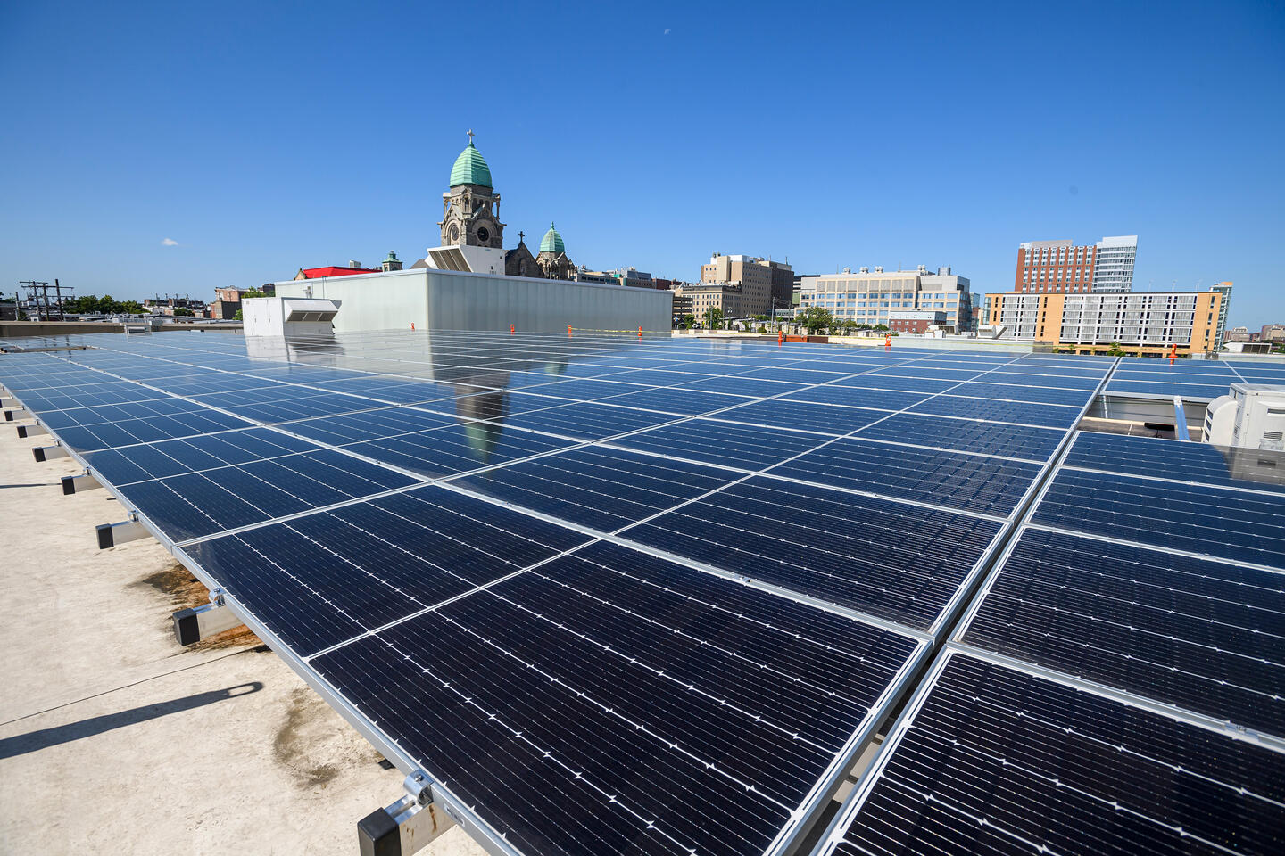 An array of black solar panels are seen on a roof in the foreground, with a skyline of city buildings, including a church, rising in the background