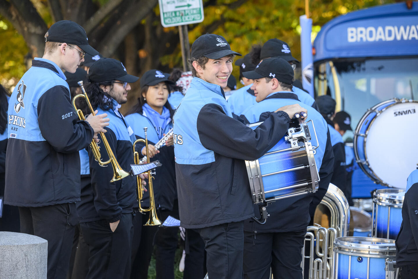 A group of Johns Hopkins pep band members talk amongst themselves in a group while in uniform, holding their instruments. One holding a drum looks back over their shoulder and smiles at the camera.