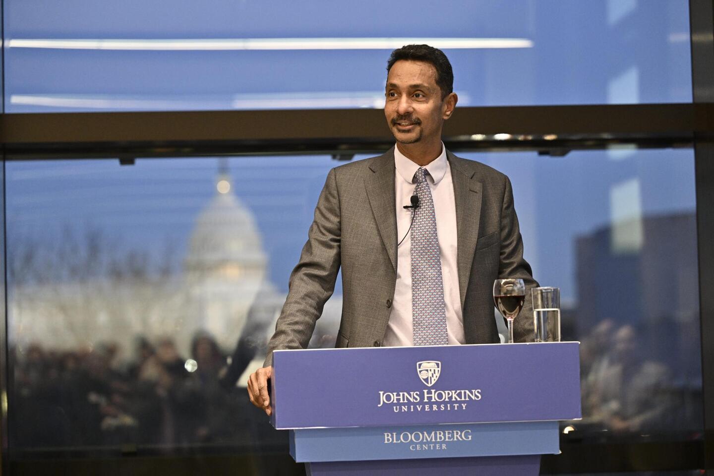 Ray Jayawardhana speaks at a Johns Hopkins-branded podium. Behind him is a window, through which the US Capitol Building can be seen.