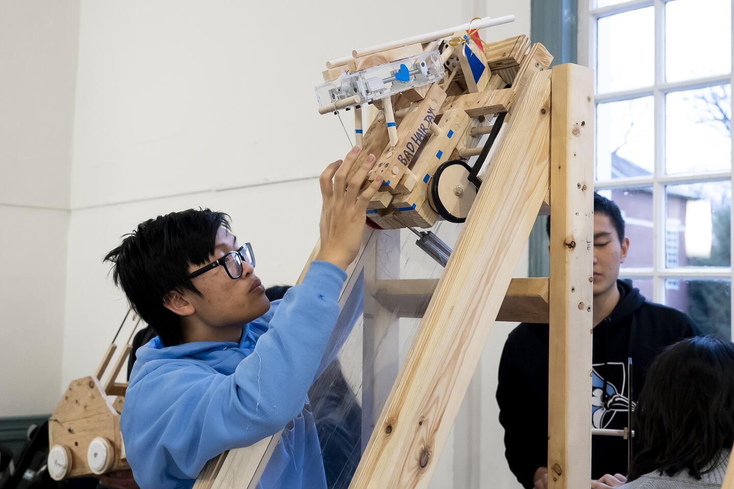 A college student sets up a homemade, wooden toy vehicle at the top of a wooden ramp.