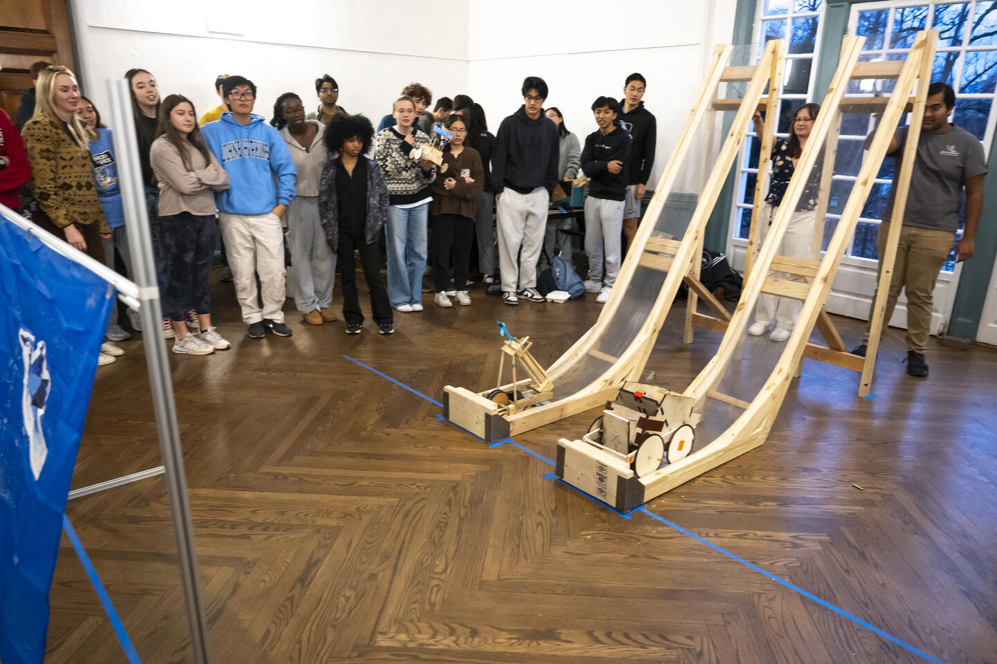 Students are gathered around two wooden ramps used to launch their self-designed vehicles