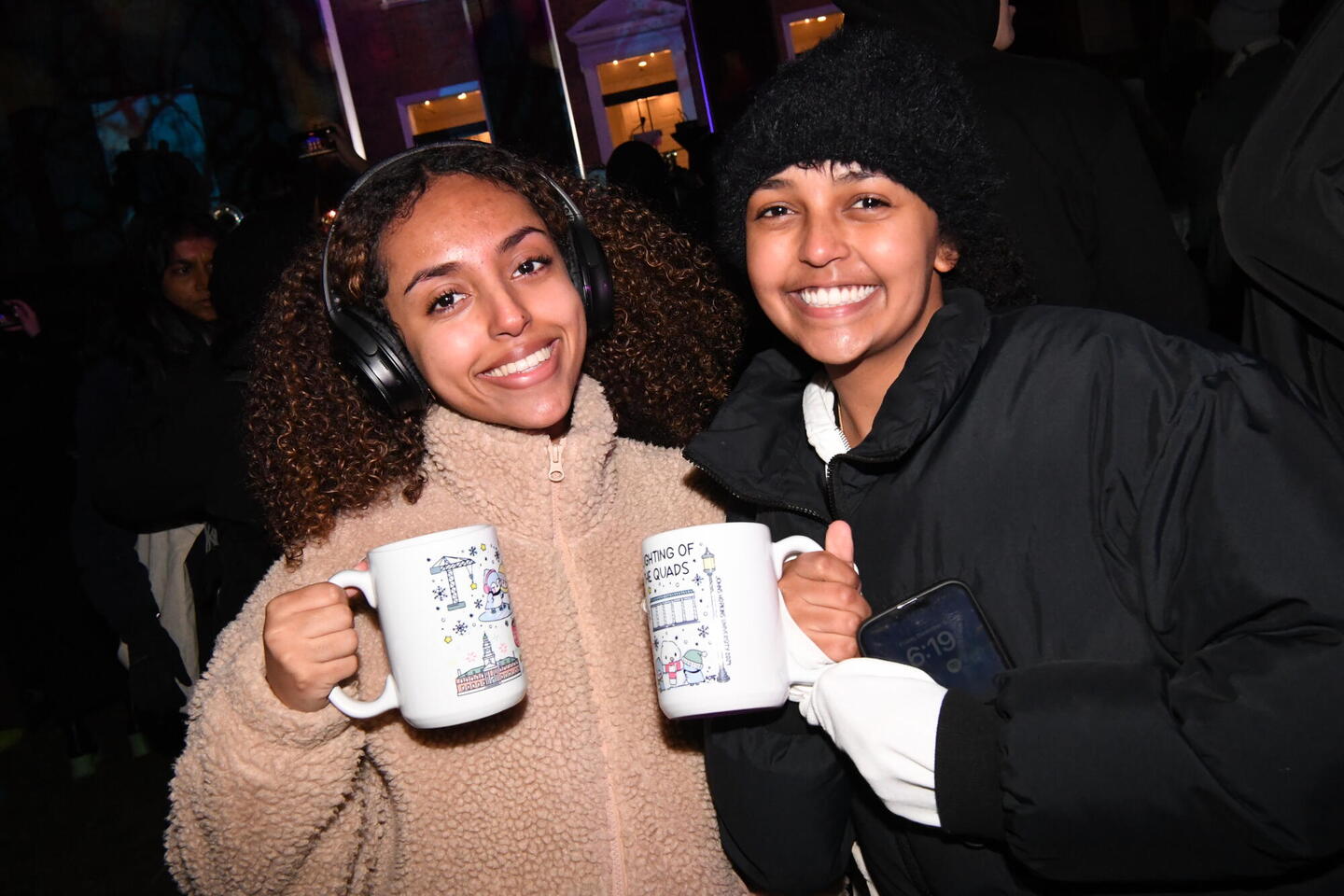 Two college students in winter clothes smile for the camera while holding up 