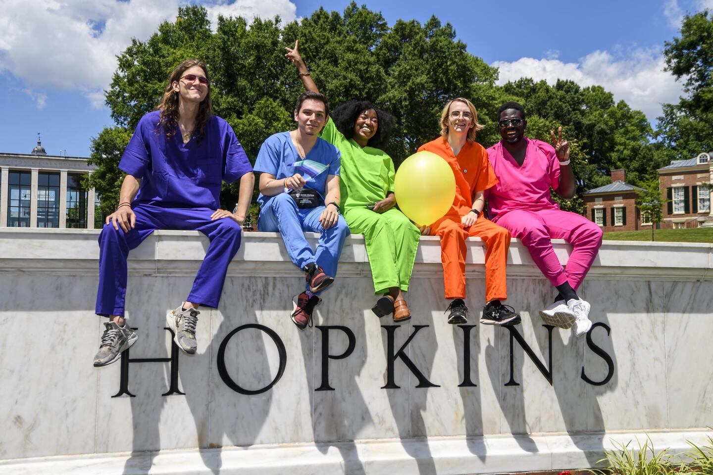 Five adults in colorful scrubs sit in rainbow order atop the Johns Hopkins sign on Homewood campus.