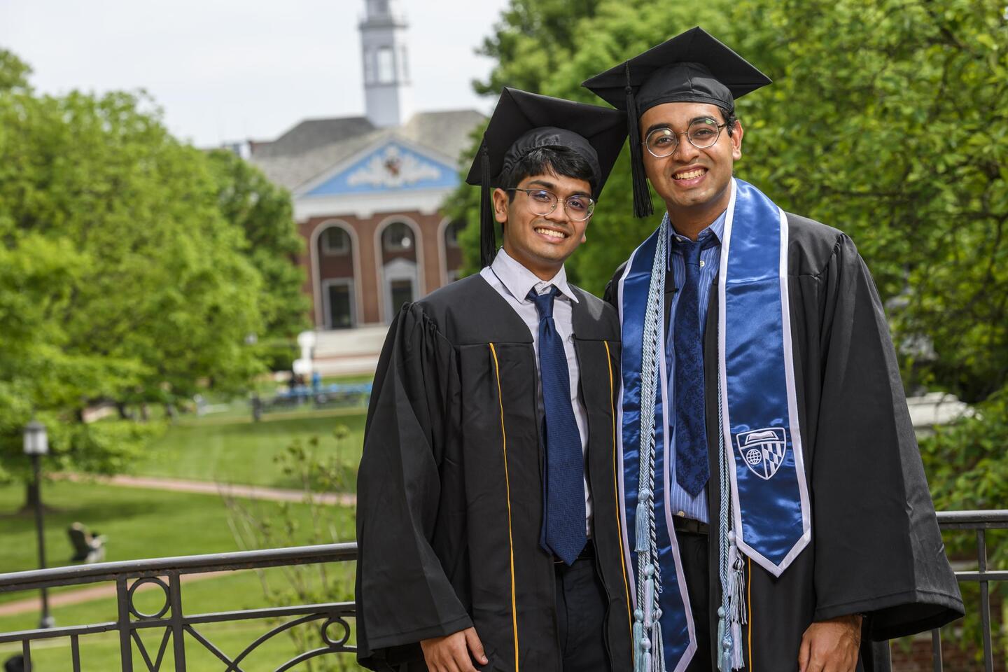 Two Johns Hopkins students in graduation robes and caps smile for a photo. In the background is Shriver Hall.