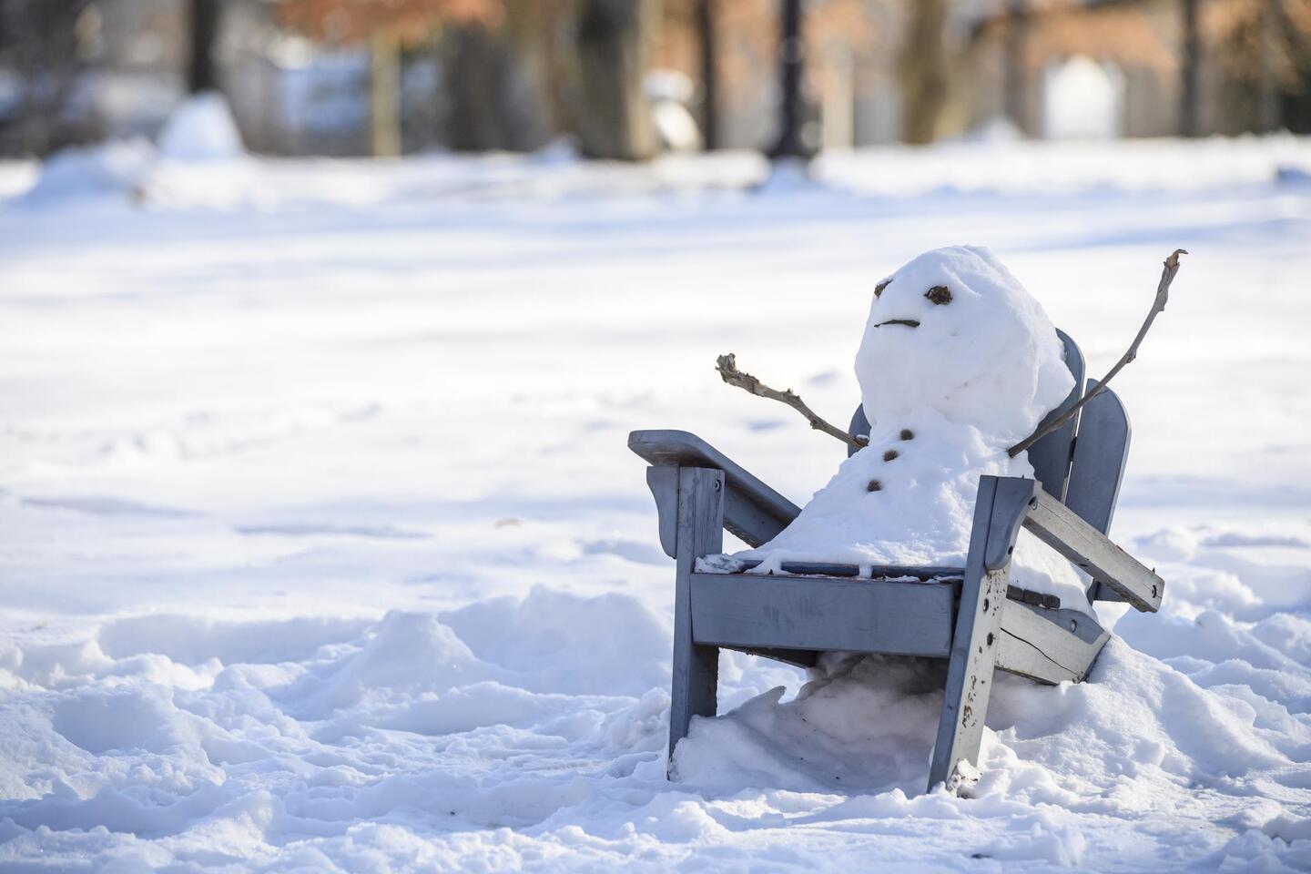 A snowman leans back on an Adirondack chair.