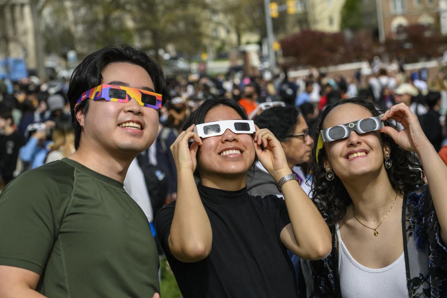 Three young adults stand in the middle of a crowd, looking up at the sky with eclipse glasses.