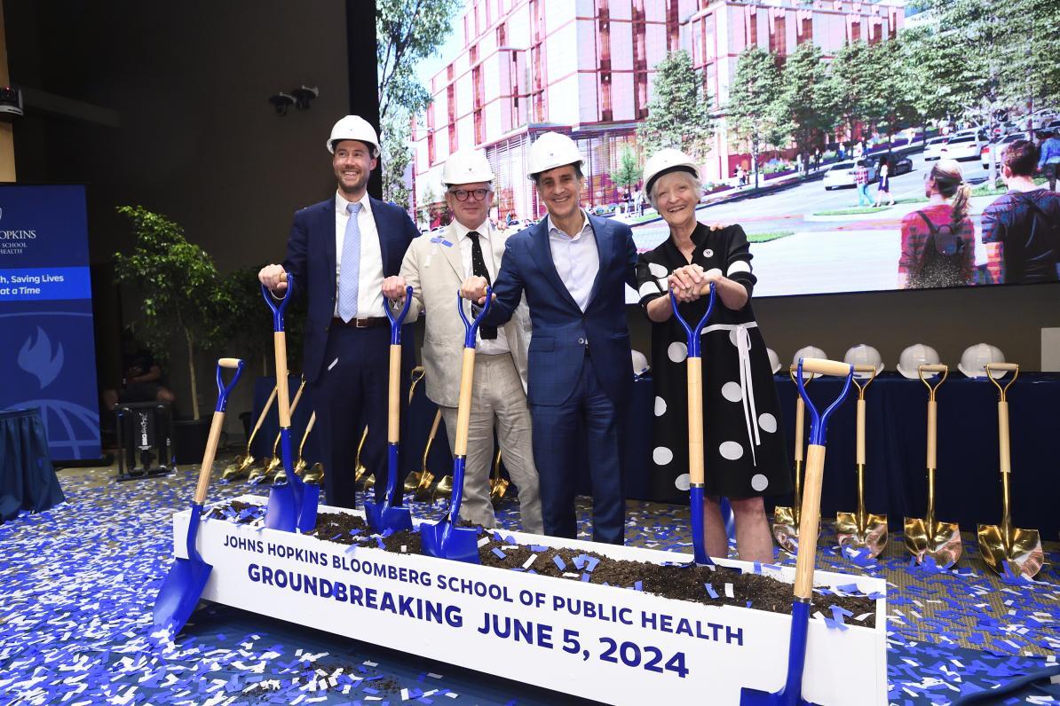 Four well-dressed adults in hard hats stand with shovels next to a trough full of dirt that reads 