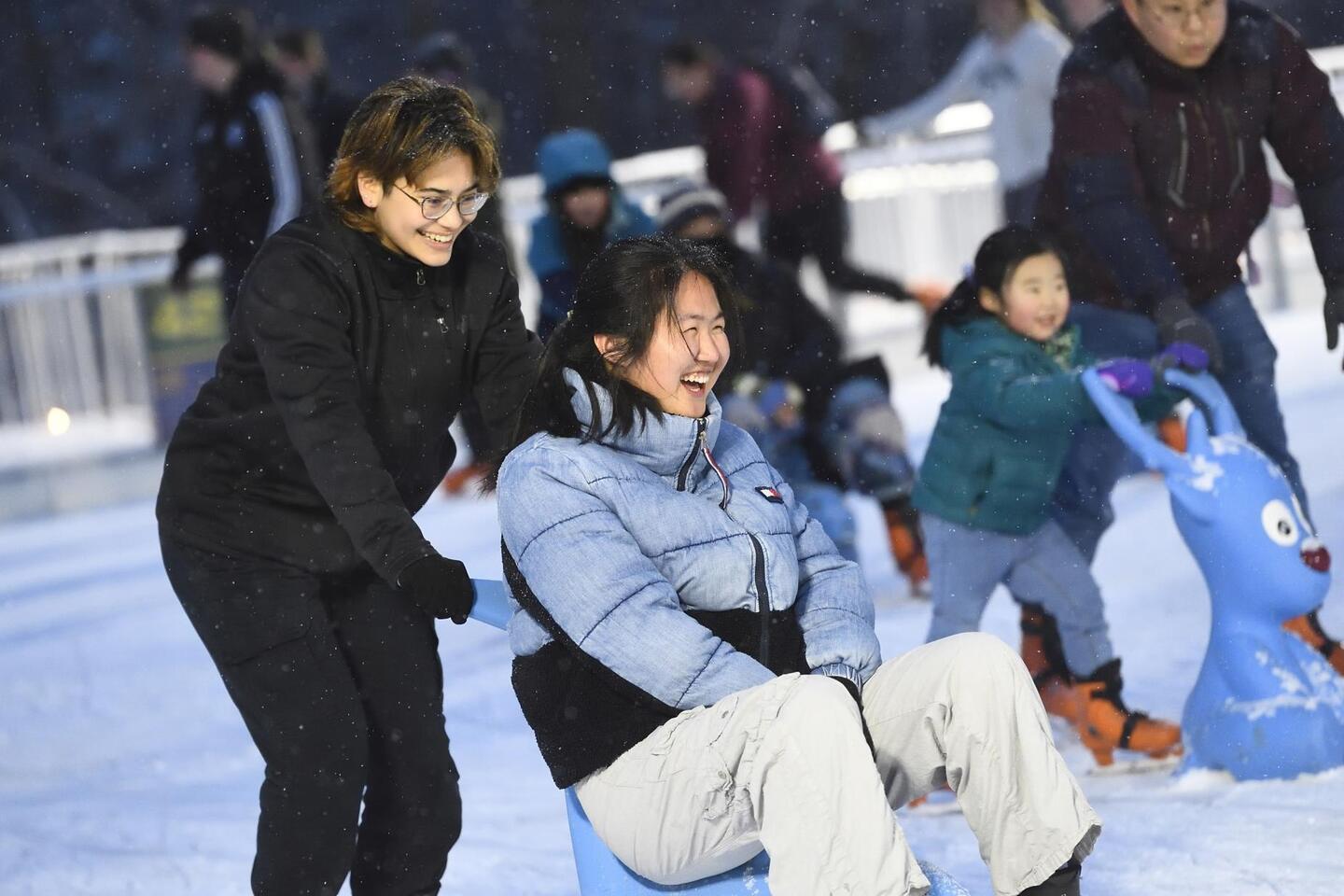 An ice skater pushes someone on chair-like sled across an ice rink.