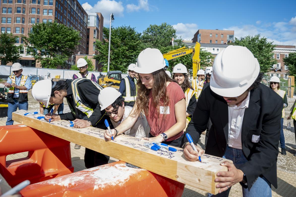 Several adults wearing hard hats sign their names on a wooden beam.