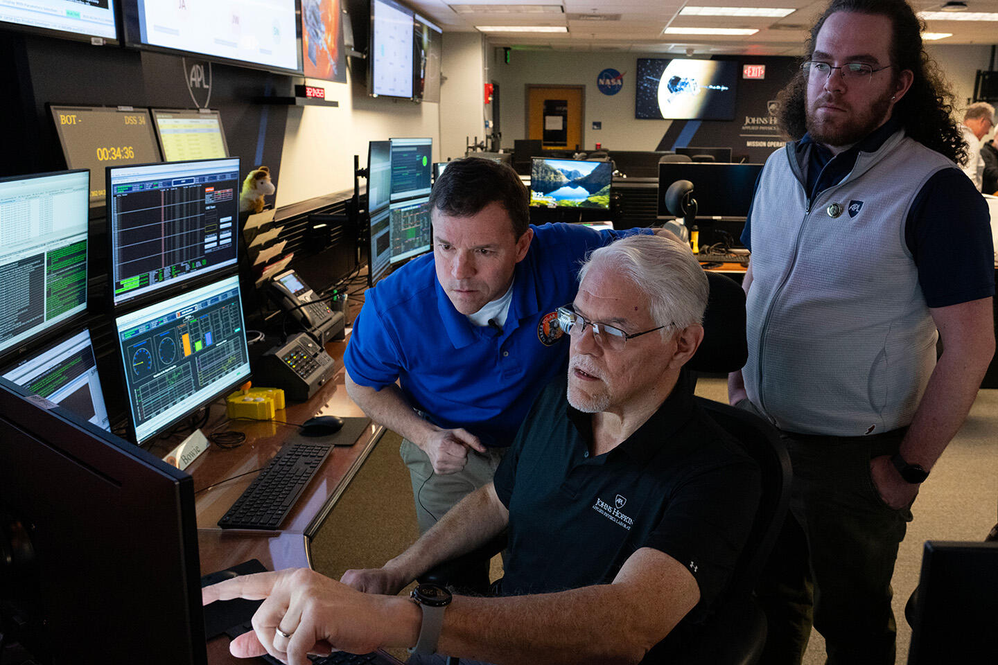 Three operators from the Parker Solar Probe mission are positioned around a desk filled with multiple computer monitors. One individual is gesturing at a screen, conversing about its details with the other two.