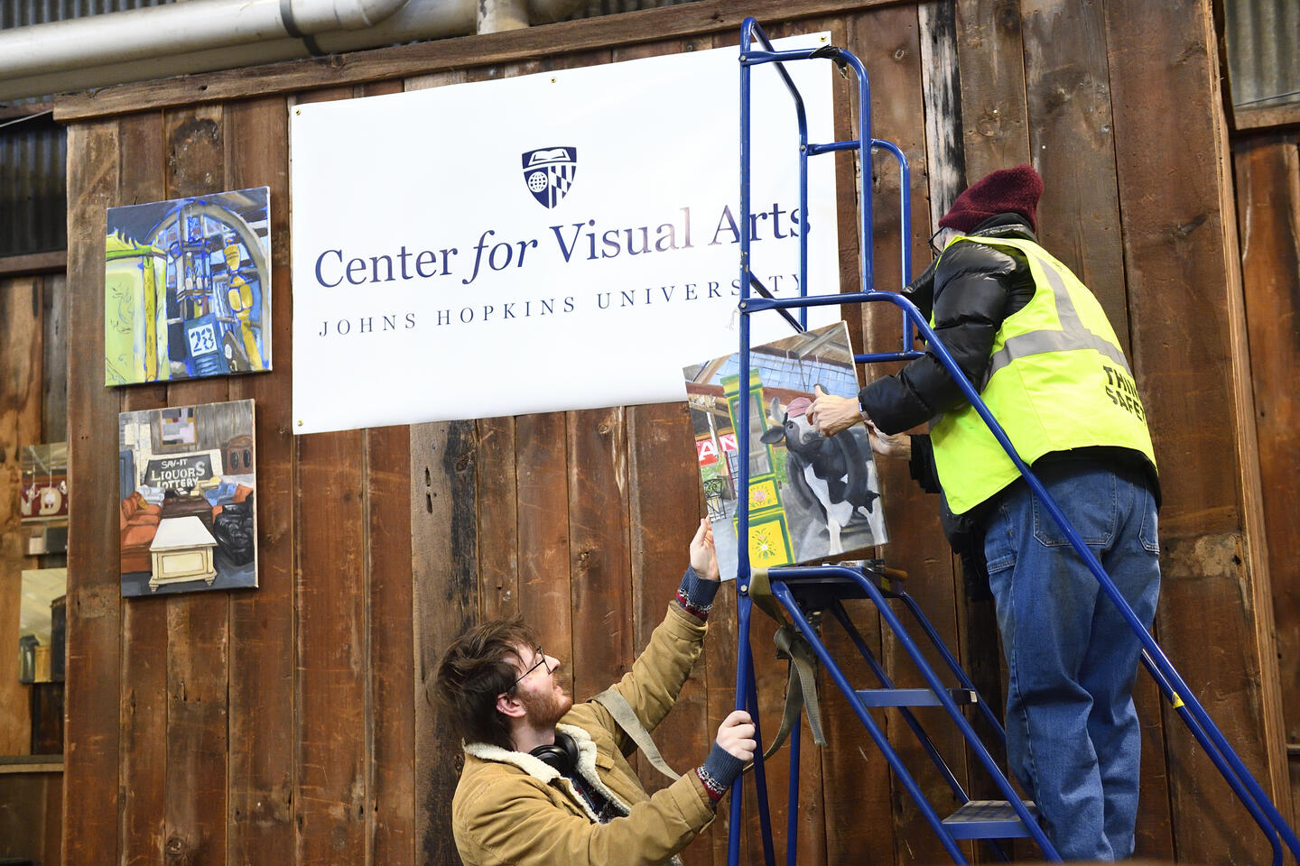 An adult hands off a painting to someone standing atop a ladder. Behind them hangs a sign that reads 