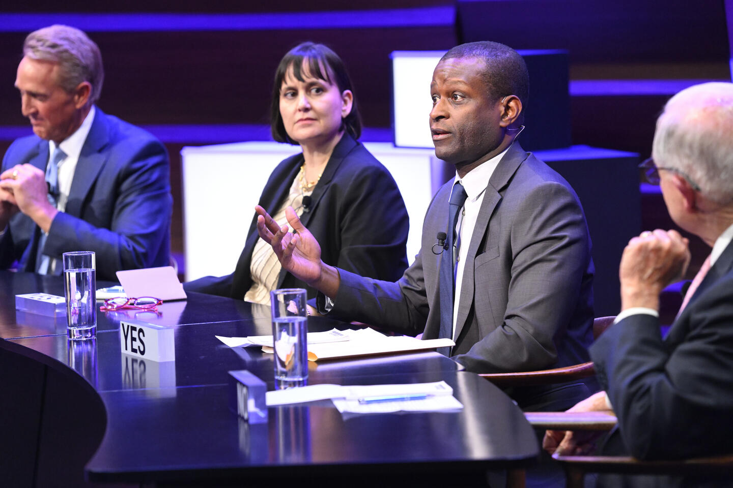 Four adults in suits sit behind a table. One in the middle talks to an audience that's out of frame.