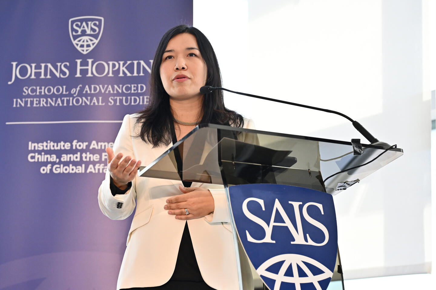 A woman in a white blazer speaks from a podium adorned with the Johns Hopkins SAIS logo