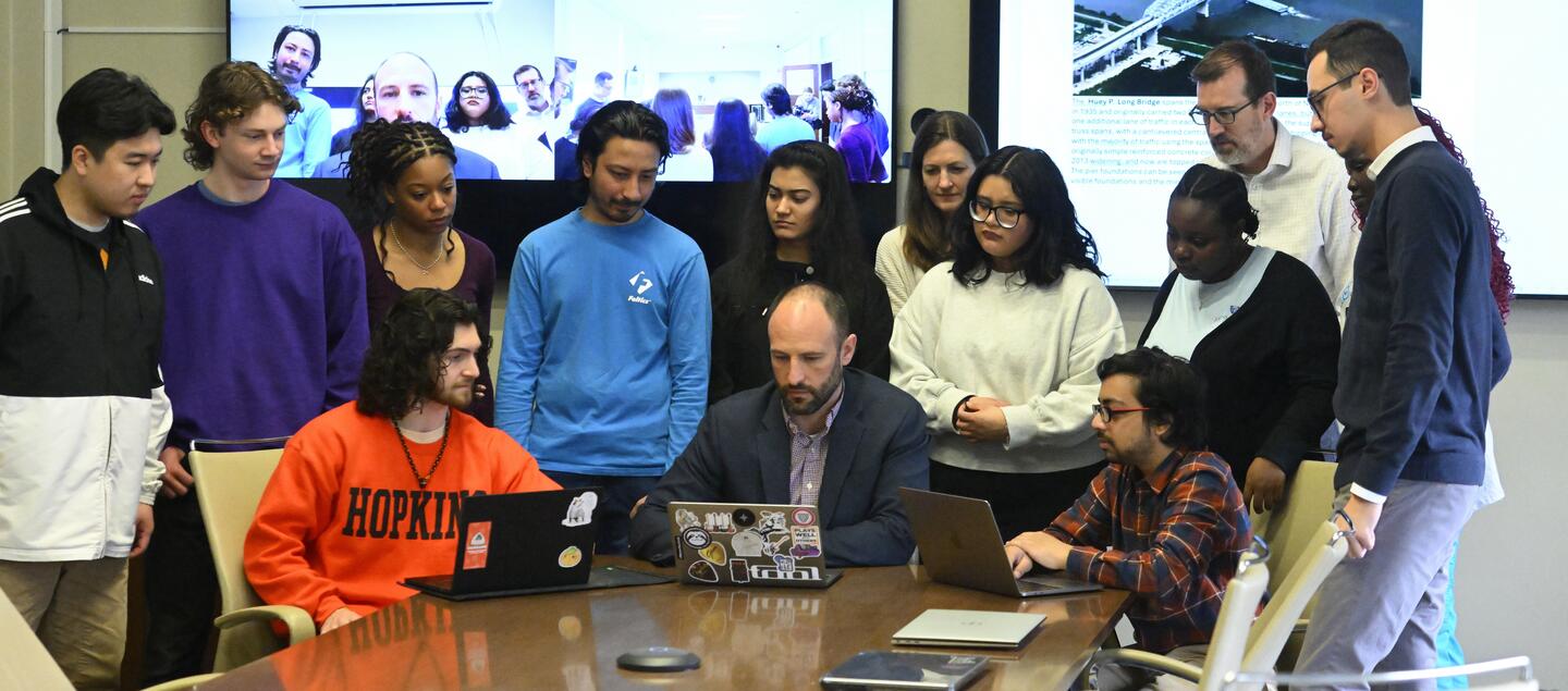 A large group of people look at a laptop screen.