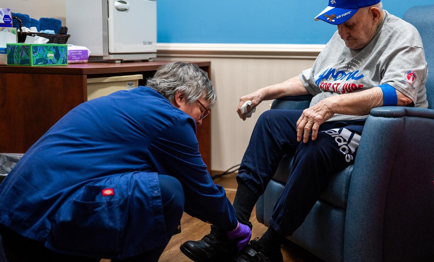 Mary Morton kneels to help Frank Squibb, who is seated in a wheelchair, with his shoes