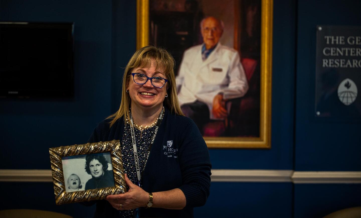 Comstock Center operations director Melissa Minotti stands in front of a portrait of George Comstock and holds a framed picture of her mother holding her as a baby