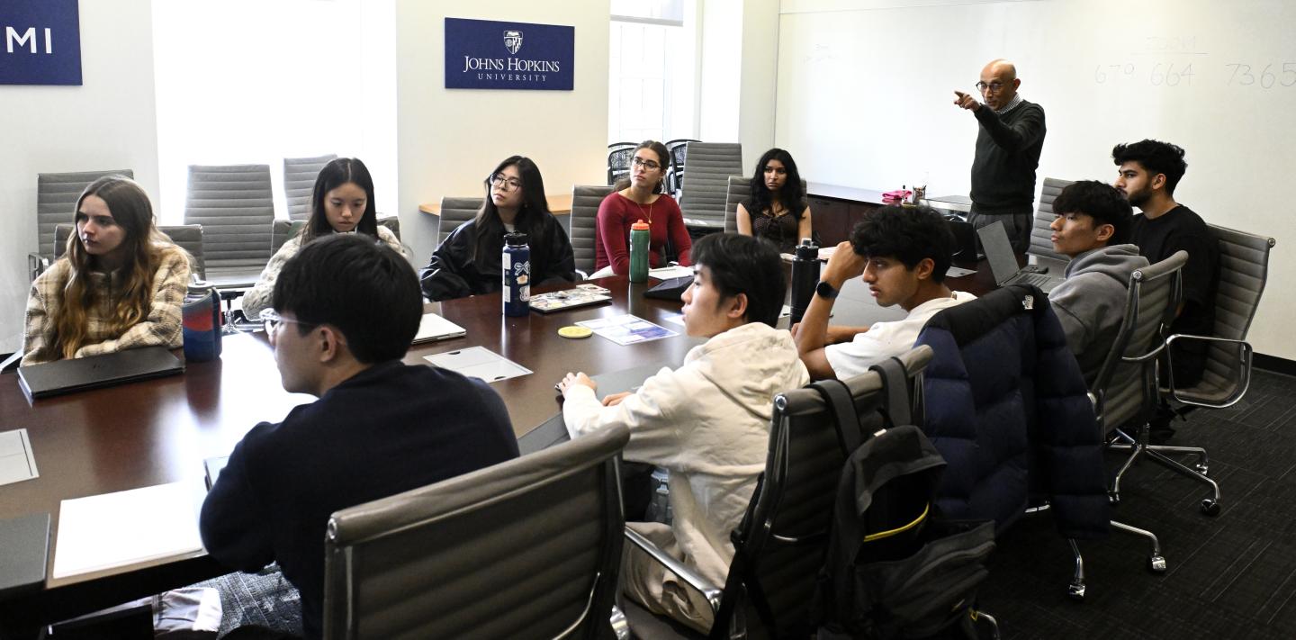 College freshmen sit on both sides of a boardroom table during a class. At the head of the table is their professor, who stands and points at something out of frame.