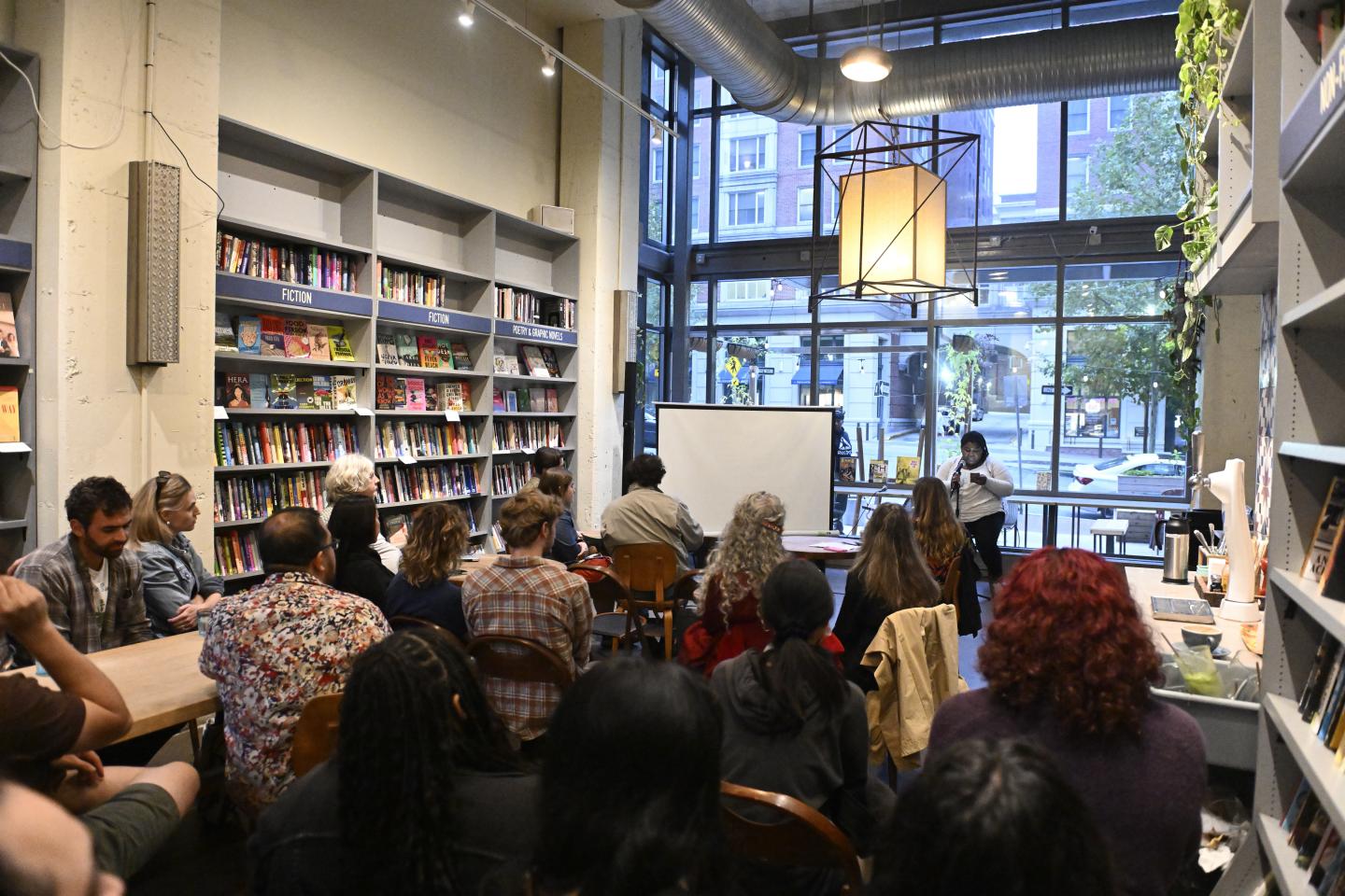 A seated person reads off their phone into a microphone to a crowd in a bookstore.
