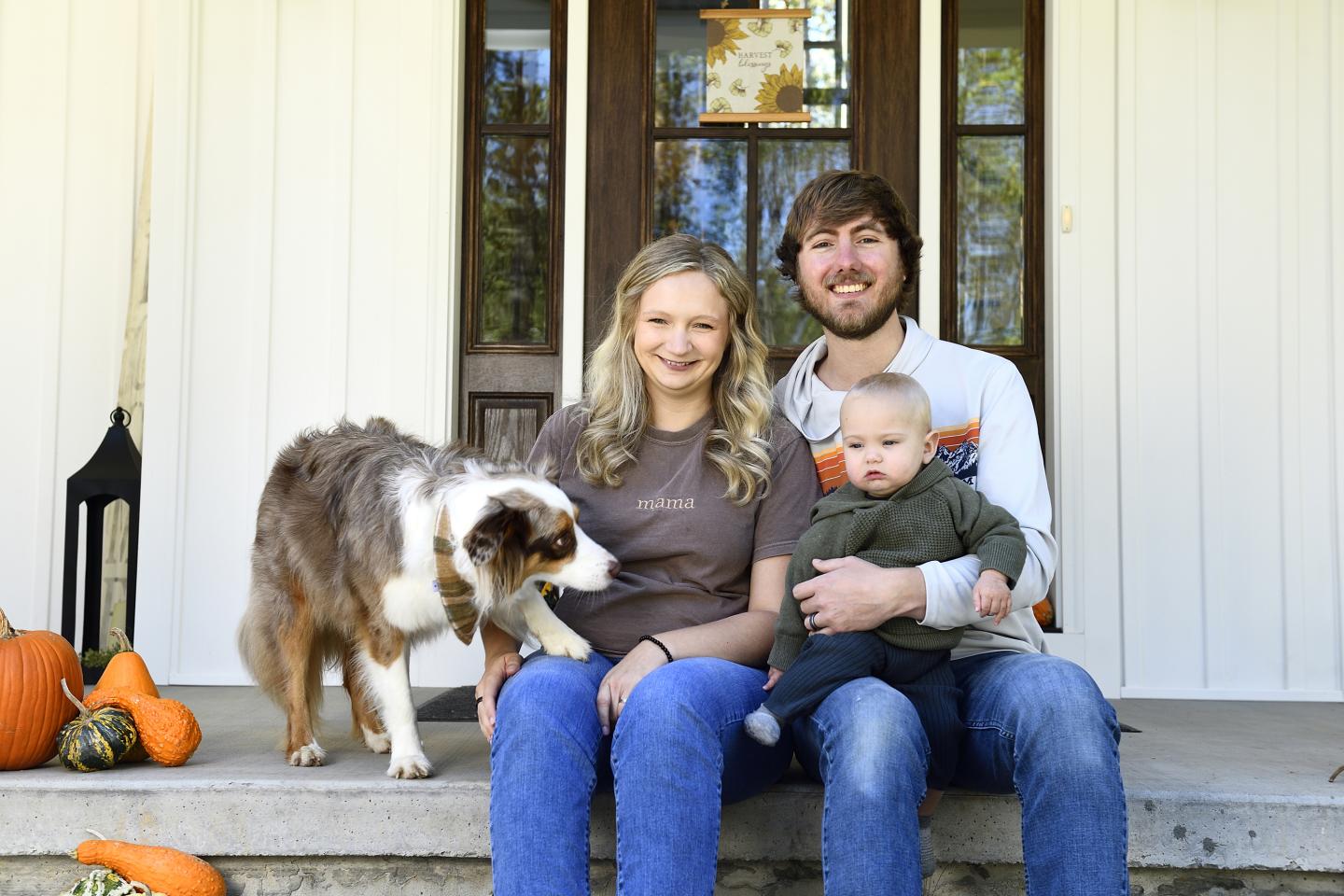 Parents Sarah and Zack Smith pose with their baby, Levi, and their dog on their front porch with seasonal gourds nearby