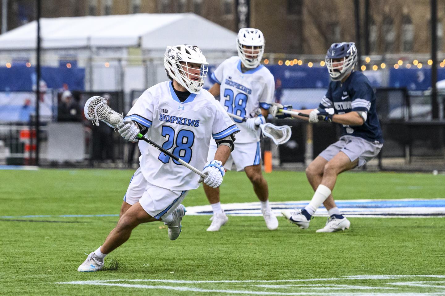 A Johns Hopkins men's lacrosse player runs with the ball. A teammate and an opponent move behind him.
