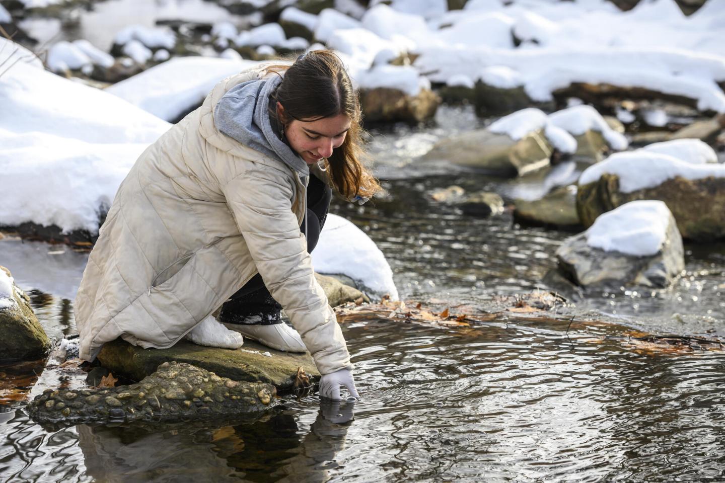 A college student crouches down to take a sample from a snowy river.