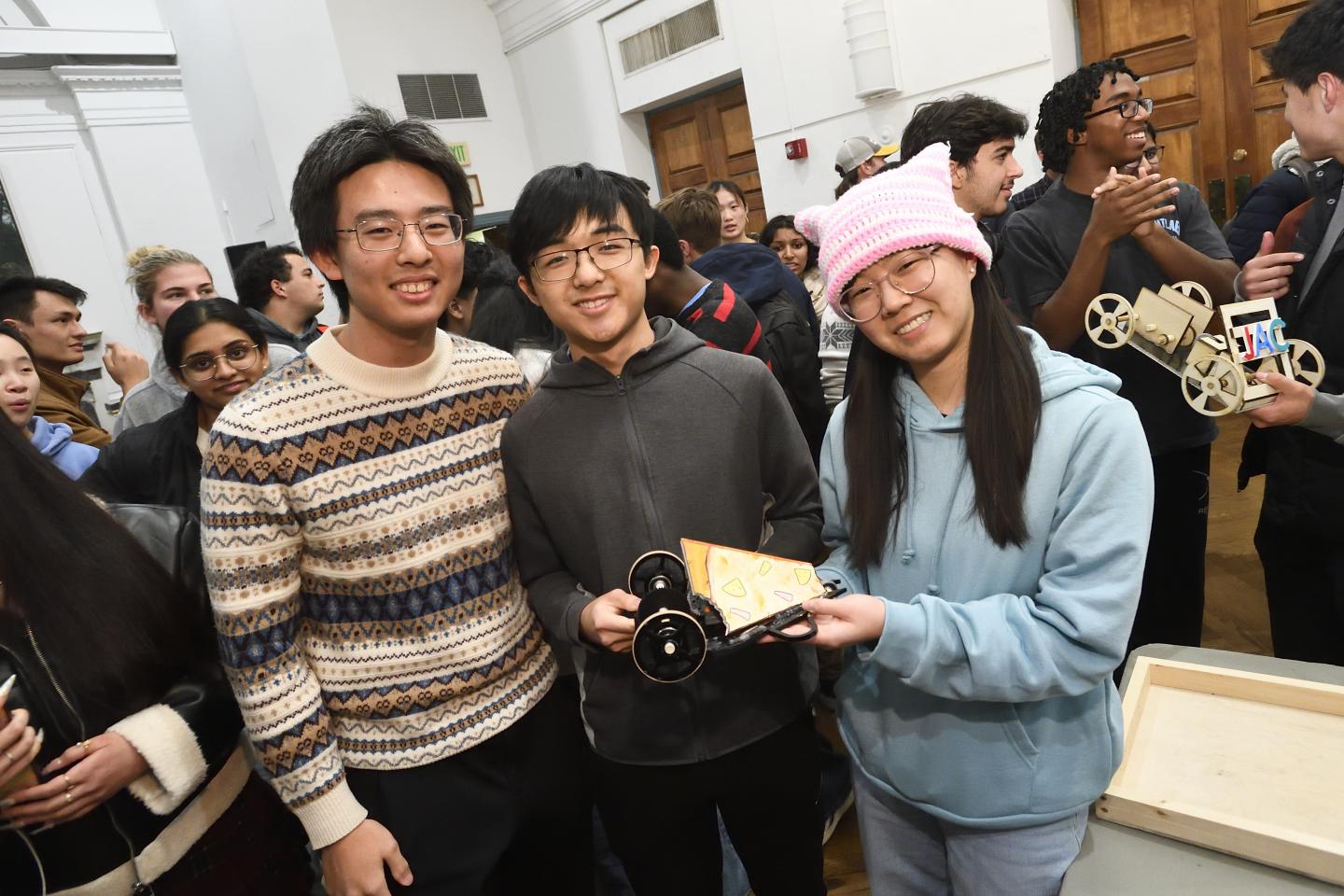 Three college students smile for the camera while holding up a homemade mechanical toy vehicle shaped like a piece of pizza.