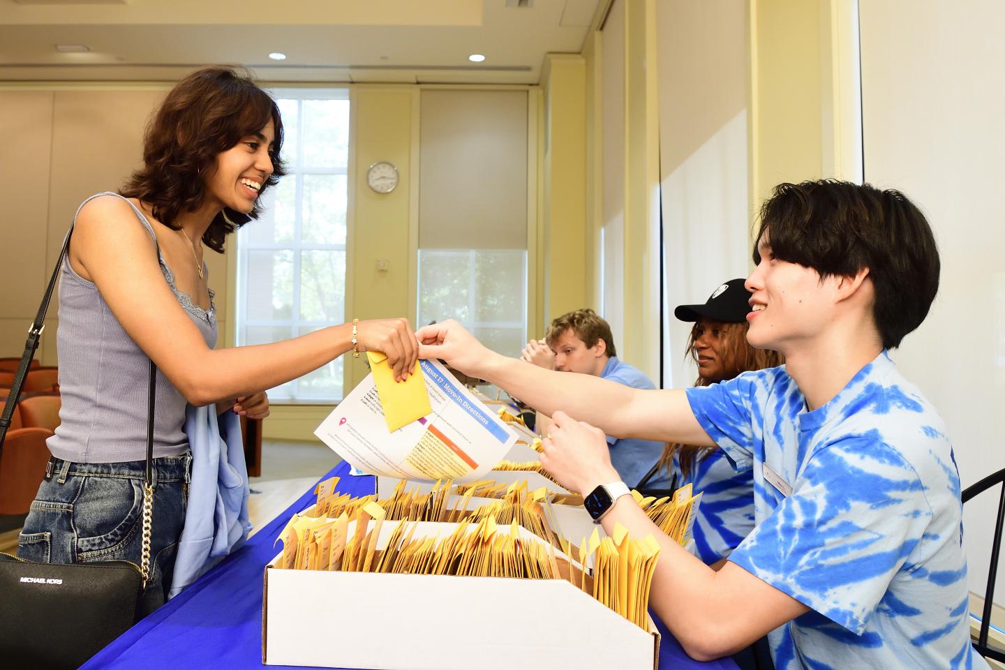 A college student hands an information packet and envelope to a first-year student.