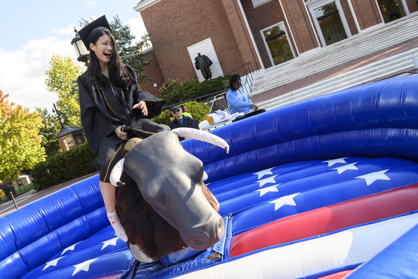 A student in a graduation cap and gown rides a mechanical bull outside.