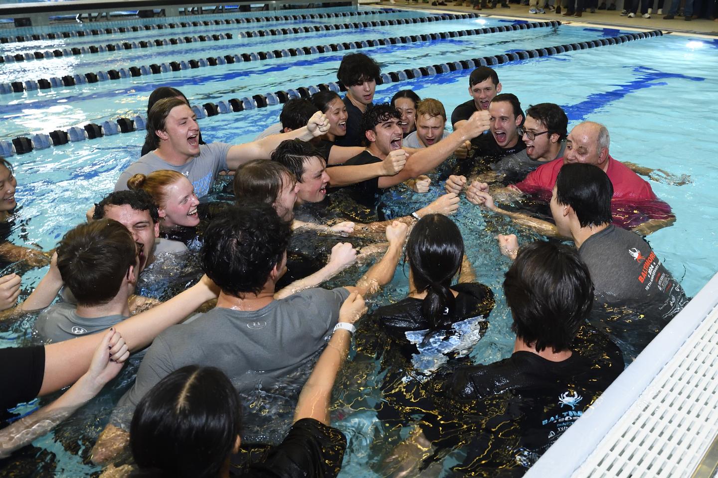 A group of people fully clothed in a swimming pool put their fists in the center for a cheer.
