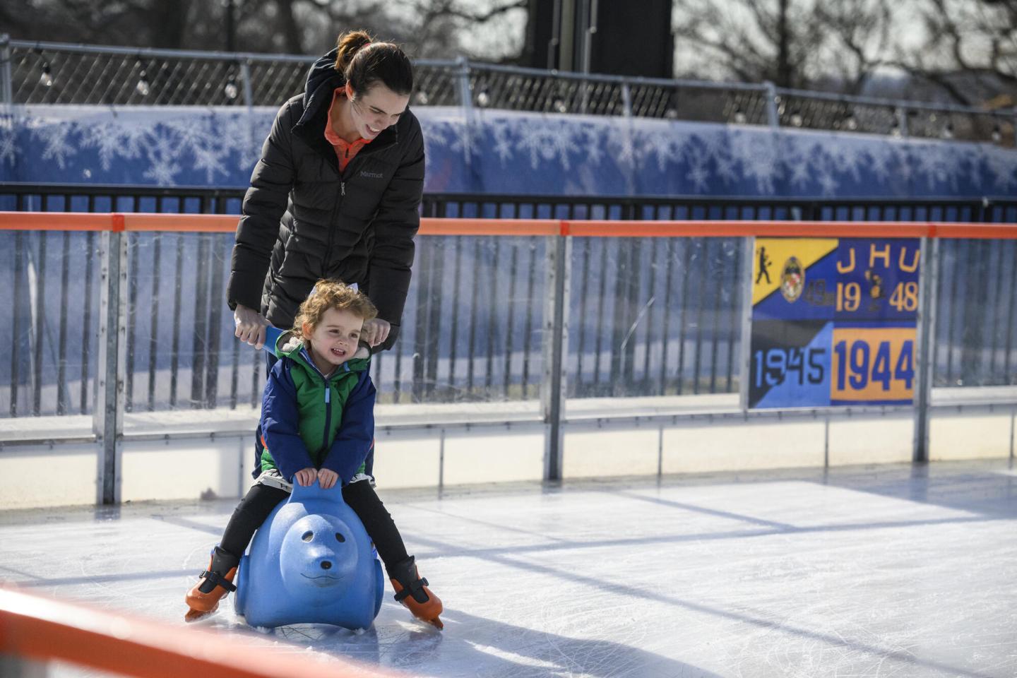 An adult pushes a child on a seal-shaped sled while skating on an ice rink.