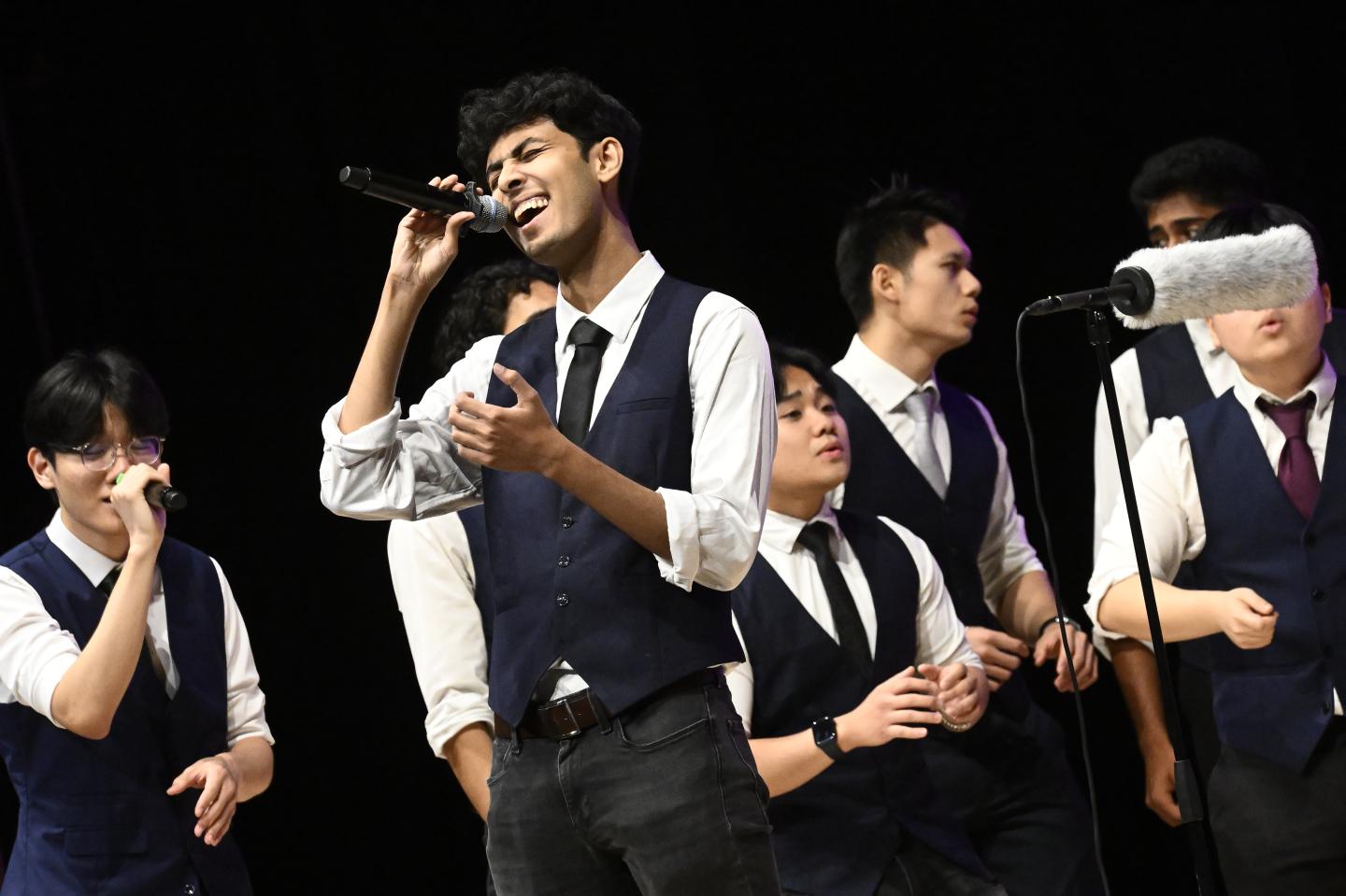 A singer belts into a microphone. More backup singers perform behind them in matching black vests and ties over white button-downs.