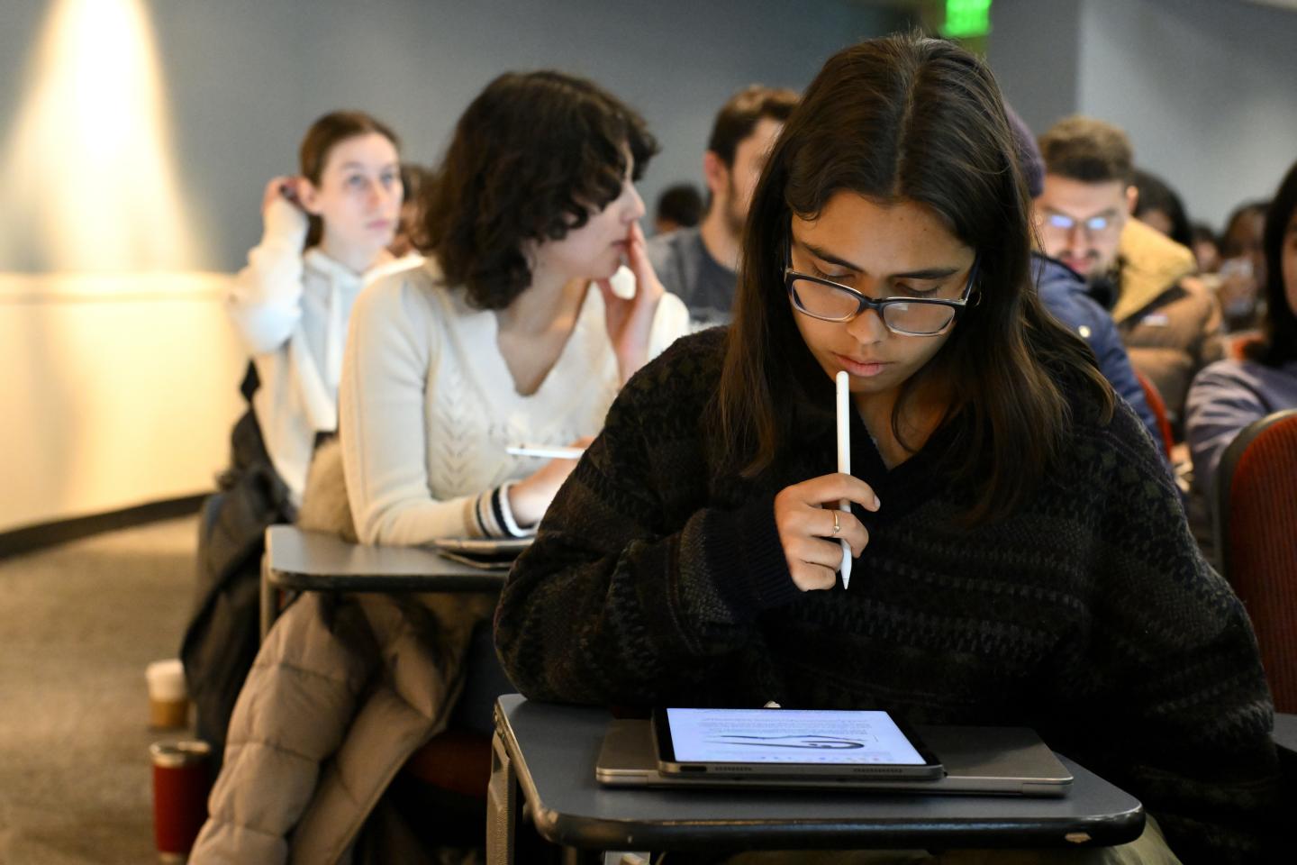 A classroom of college students sit, waiting for class to start. The student in front brings their stylus to their lips, reading something on their tablet.
