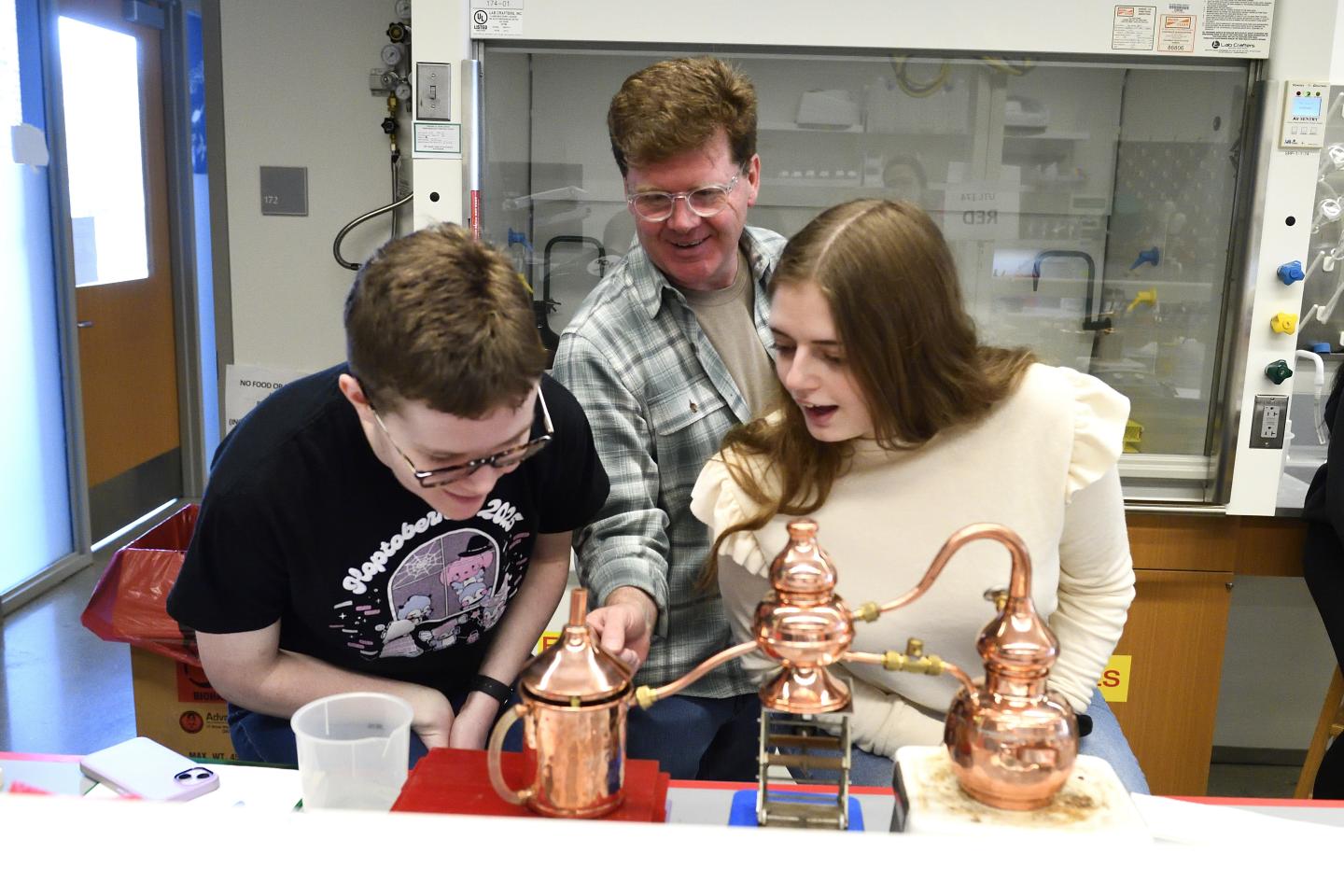 Three people react with delight as a small copper still produces alcohol.