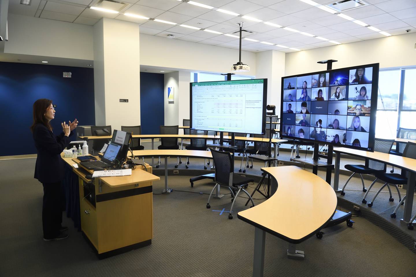 A woman in an empty seminar room teaches in front of a large digital screen of students