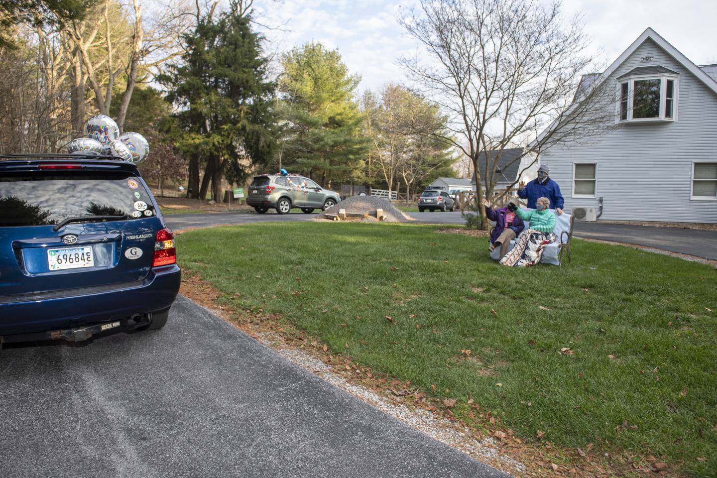 A line of cars with balloons drive past two women who are seated and one man standing