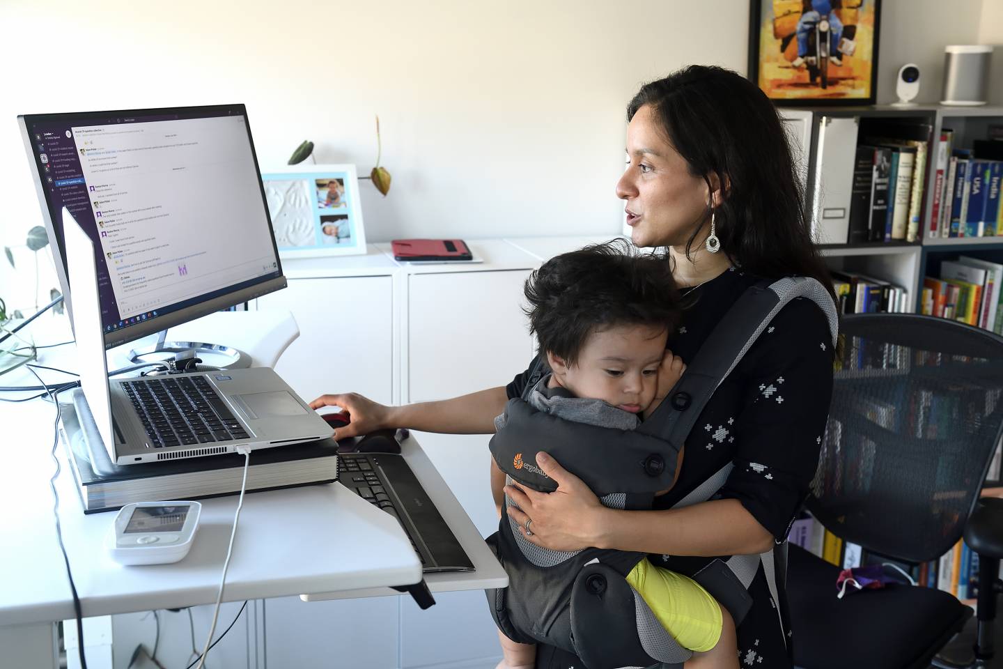 A woman works at a computer with a baby in a carrier strapped to her chest
