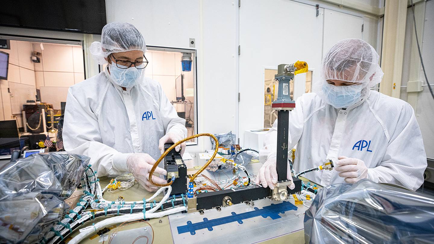 Two scientists wearing protective gear work side-by-side on a spacecraft