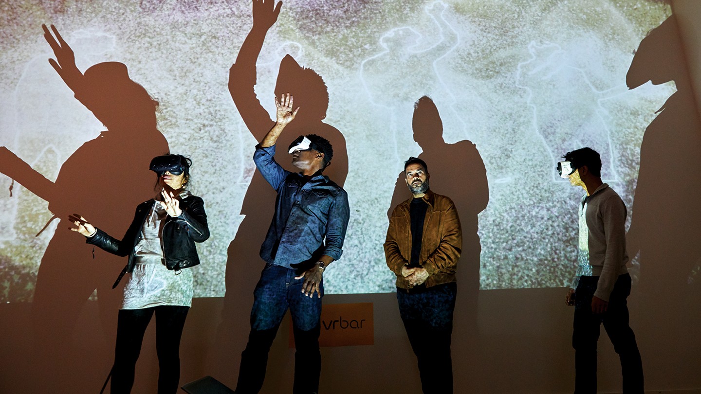 Three people wear VR goggles while a fourth looks on Three people wear VR goggles while a fourth looks on
