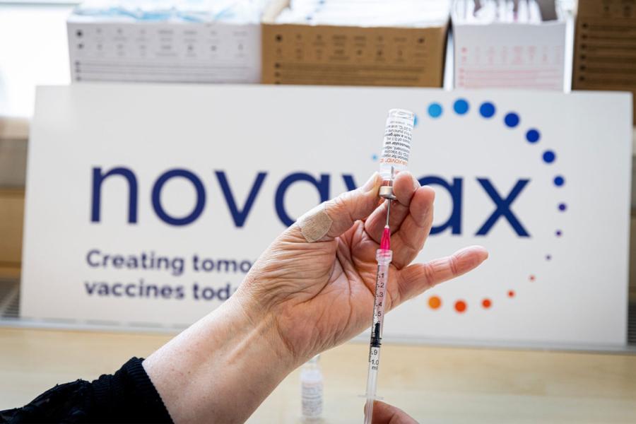 A person loads a syringe from a glass vial containing the Novavax vaccine while a promotional sign in the background reads 'Novavax'
