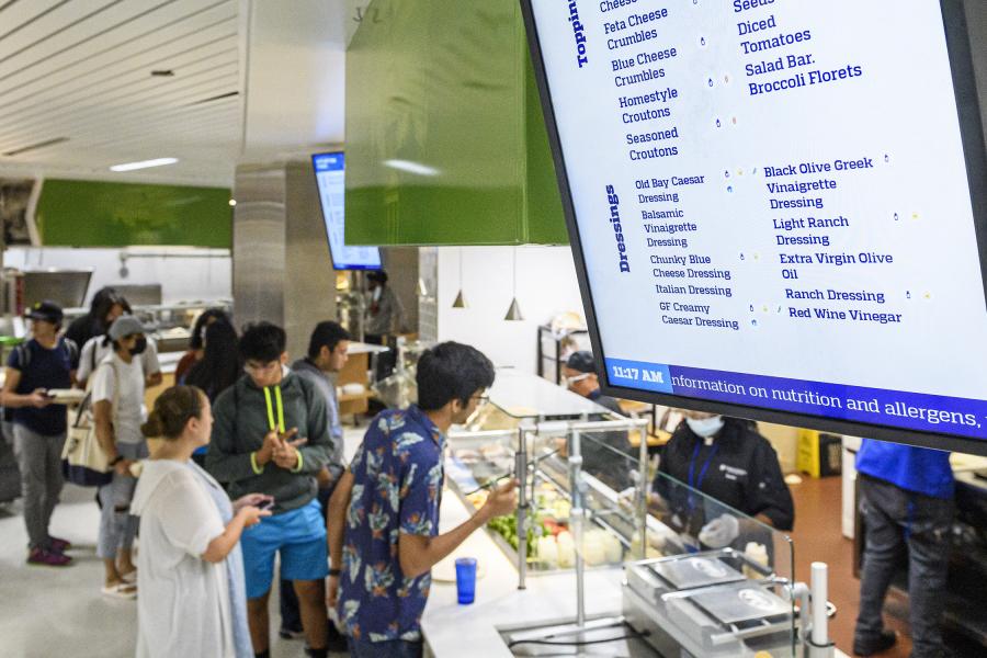 A digital screen displays a menu while students order food in the background